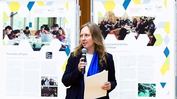 A woman with a microphone and sheets of papers in her hand in front of two posters