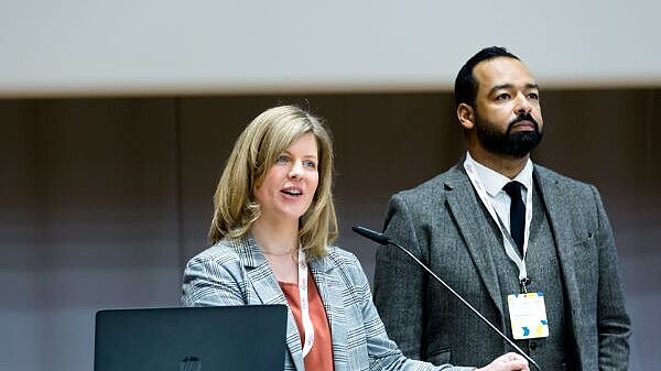 A woman and a man standing on a podium, the woman speaking in the microphone