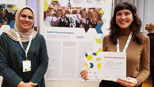 Two women standing in front of poster, one holding a certificate in her hands